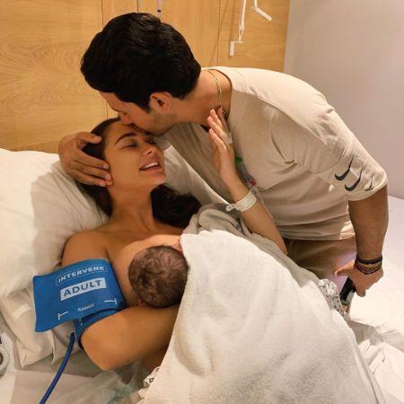 Amy Jackson breastfeeding her newborn baby lying on a hospital bed while her husband George Panayiotou kisses her on the forehead.